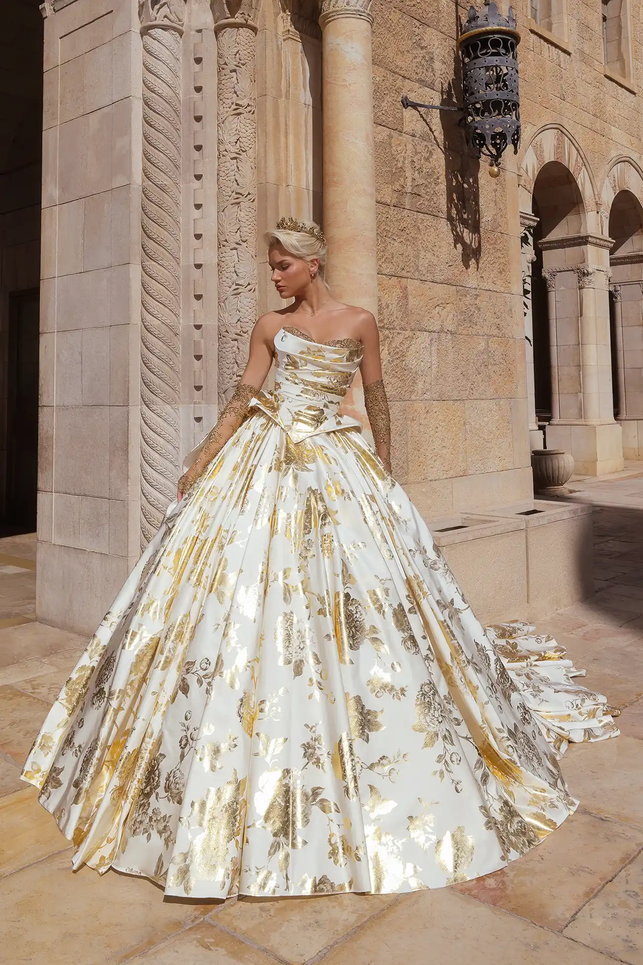 Strapless ballgown in matte satin printed with gold foil peonies, with signature Pnina bow, draped basque waist bodice and gold crystal embellished bust detail. Shown with matching gold crystal metallic chantilly lace gloves.