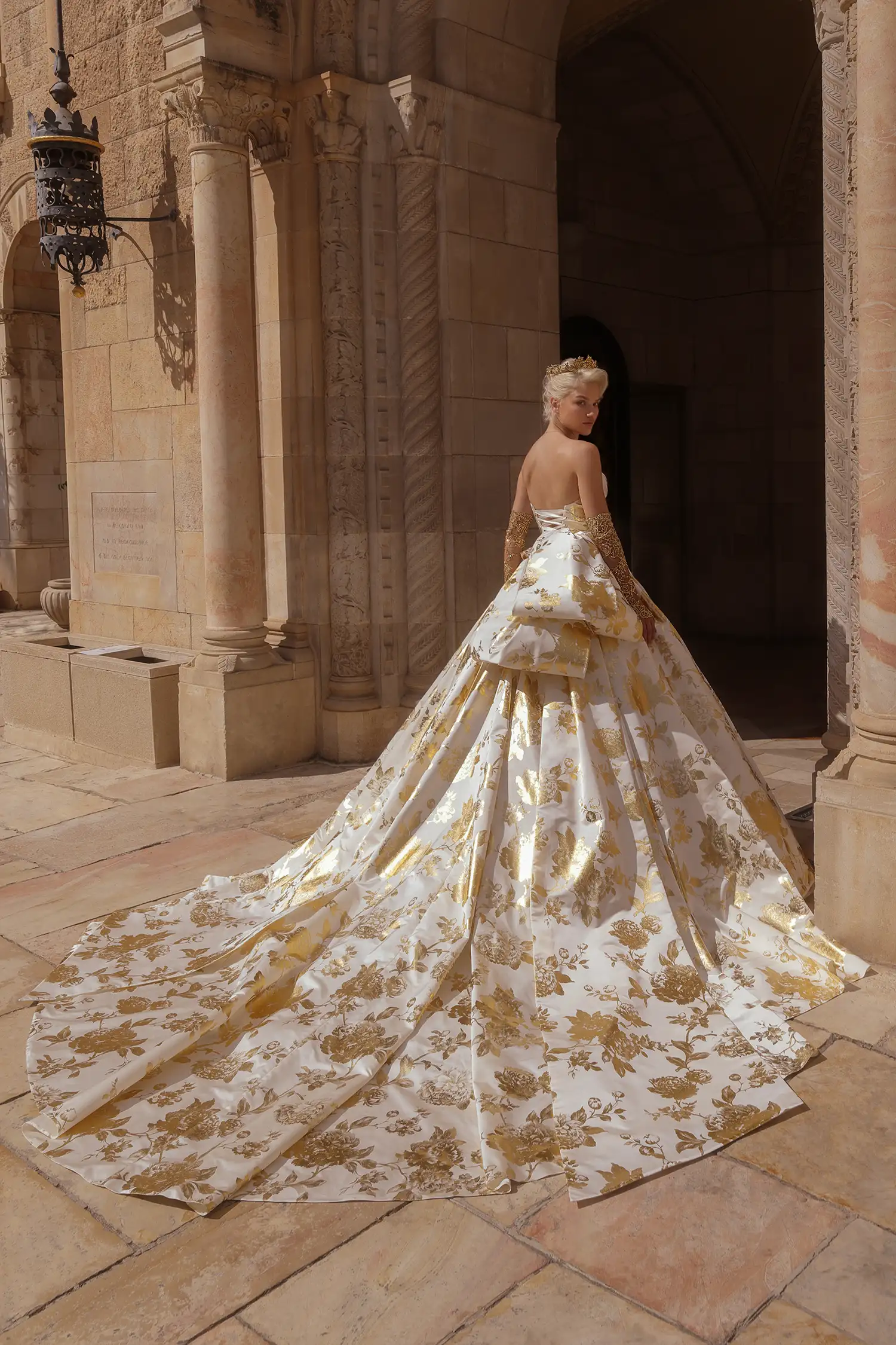 Strapless ballgown in matte satin printed with gold foil peonies, with signature Pnina bow, draped basque waist bodice and gold crystal embellished bust detail. Shown with matching gold crystal metallic chantilly lace gloves.