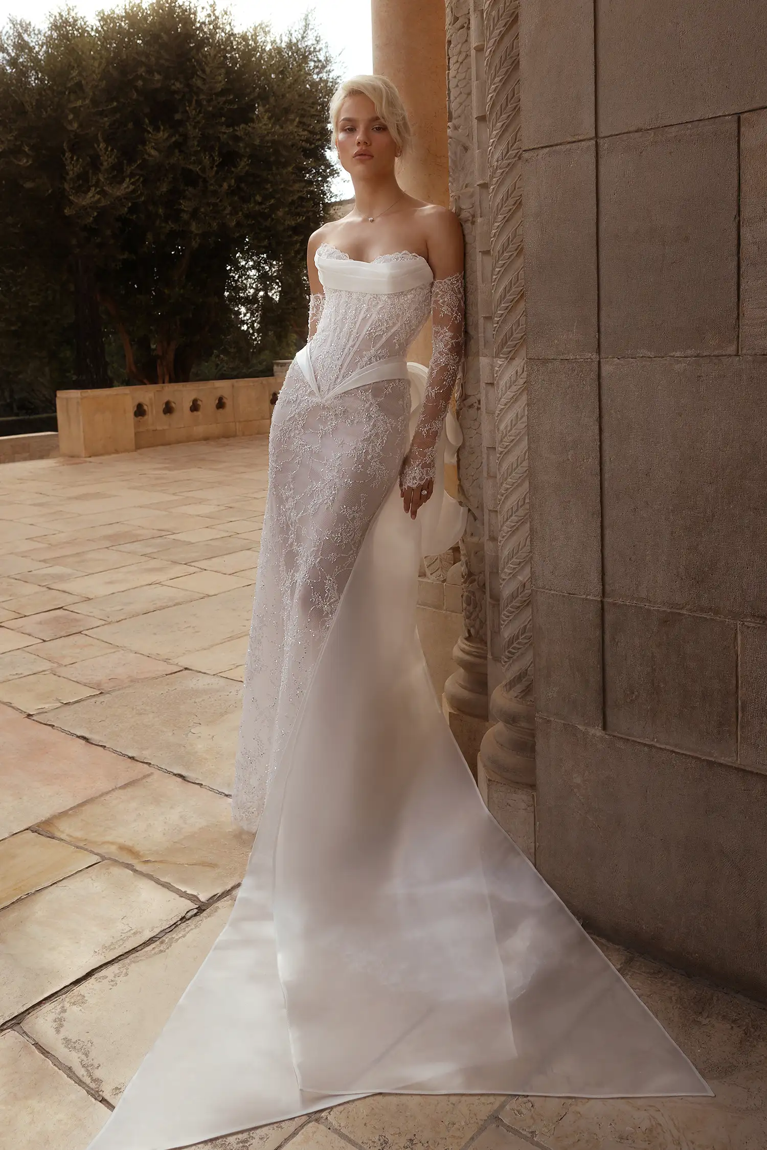 Hand-beaded chantilly lace sheath gown with high-shine liquid organza draping at neckline and basque waist and signature Pnina bow. Shown with beaded chantilly lace gauntlet gloves. 