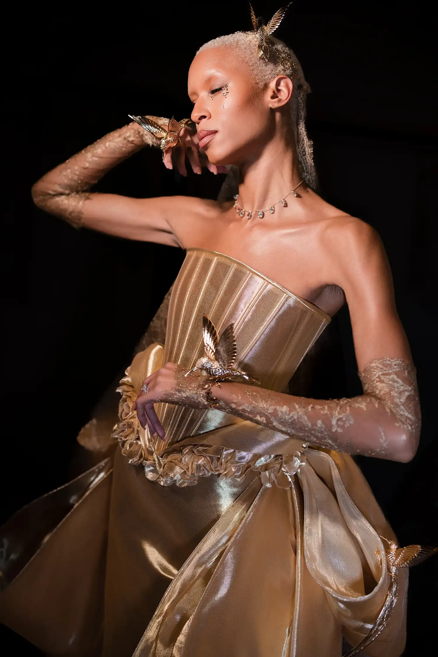 Strapless liquid gold fit-and-flare gown with basque waist accented with handmade dimensional roses, golden hummingbirds, and a large signature Pnina bow at back, shown with gold metallic chantilly lace gloves and veil.