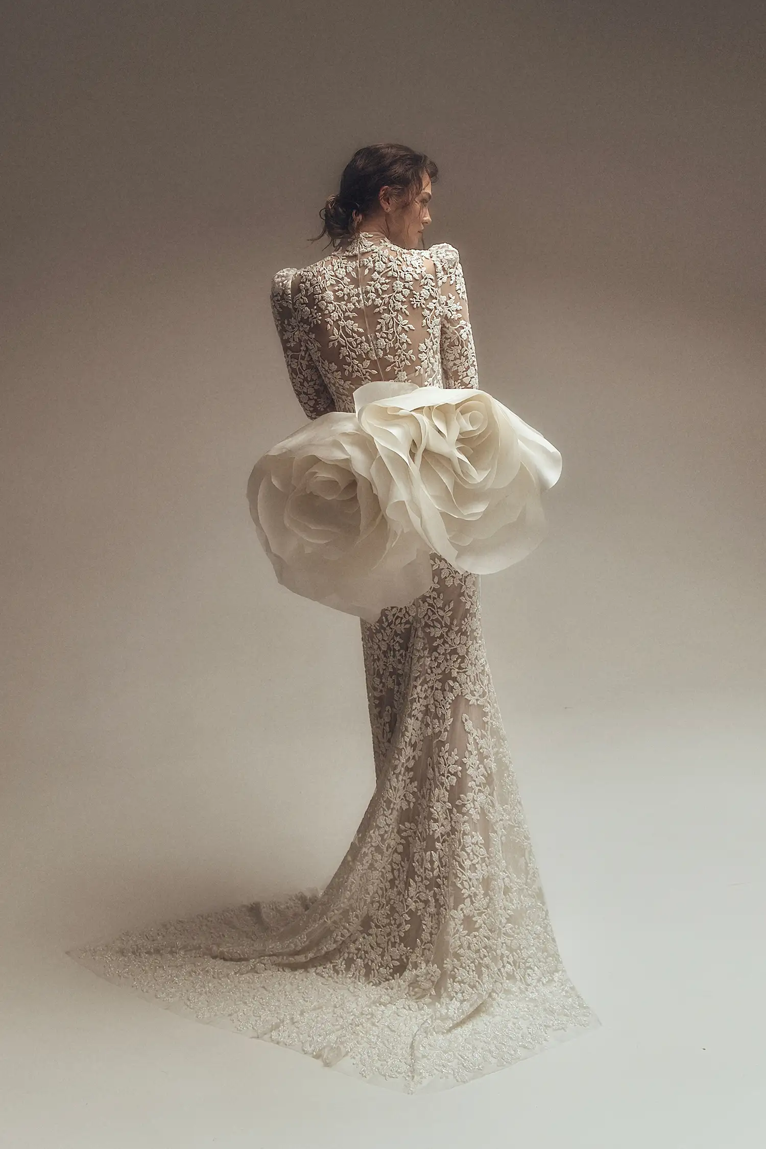 Long sleeved high neck sheath gown in embroidered lace fully hand beaded with tiny pearlized bugle beads. Shown with satin-face silk organza open overskirt with pickups and oversized handmade roses and matching hand-beaded lace mantilla veil.