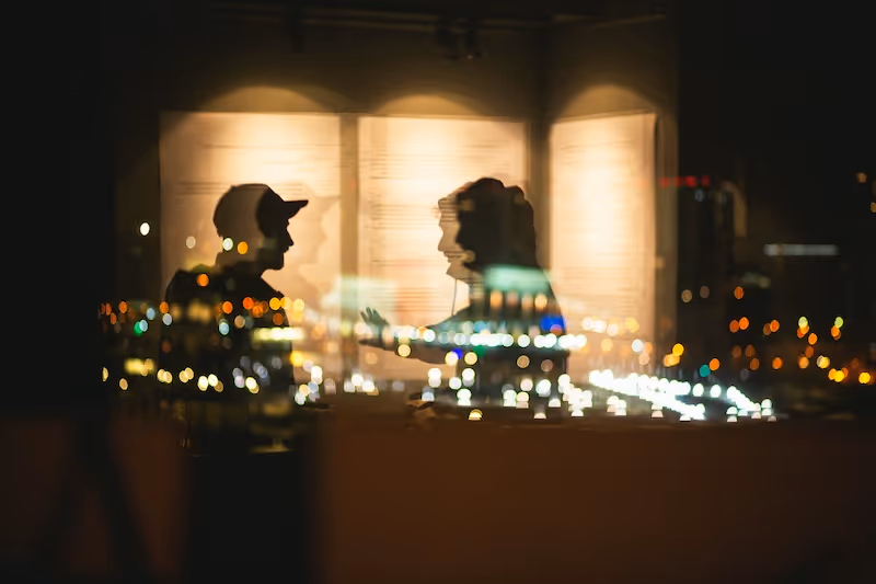 A stock photo of two people's silhouettes on glass, at night, with city lights shining through the window  