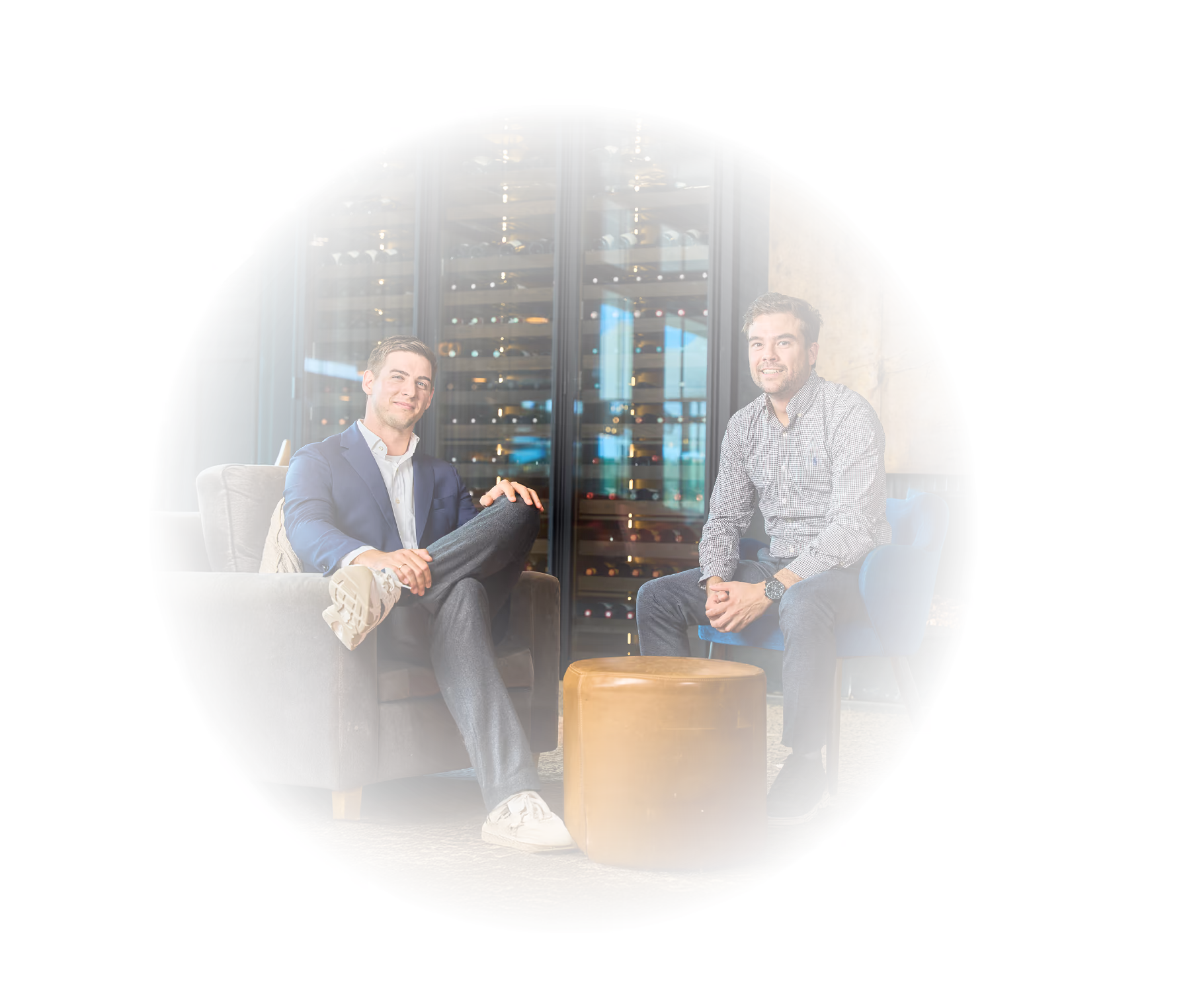 Two men sitting and smiling in a modern lounge area with a wine display behind them and a brown ottoman in front.