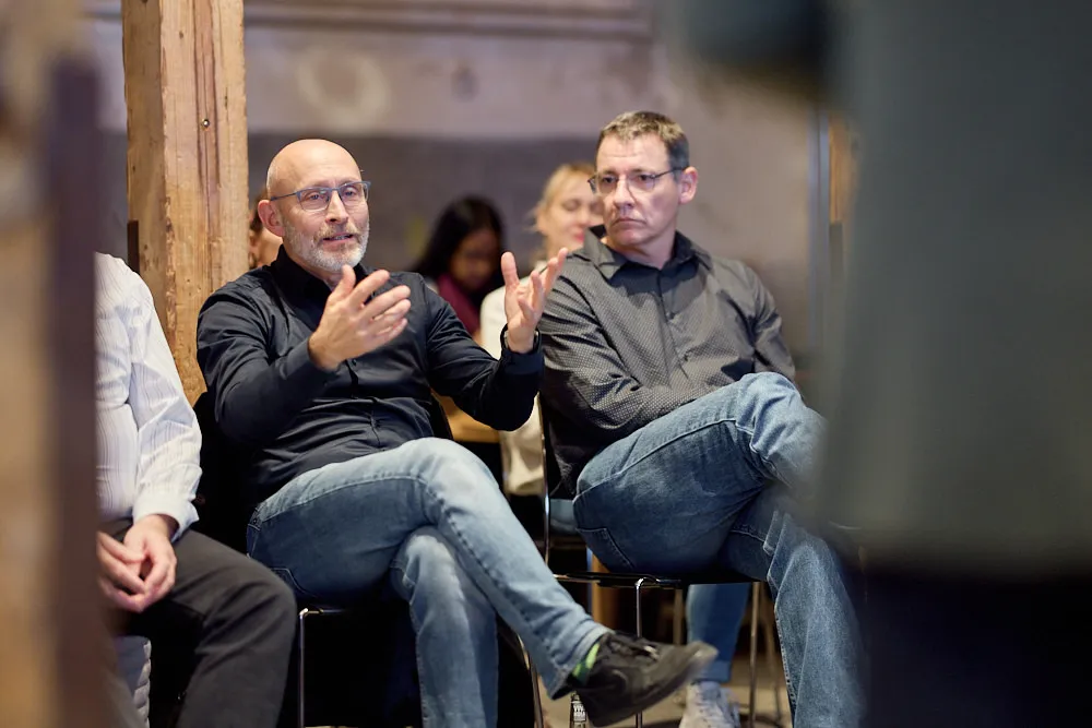 Im Fokus sind zwei Männer im Publikum. Der ältere Mann mit Bart erzählt etwas und gestikuliert mit den Händen.