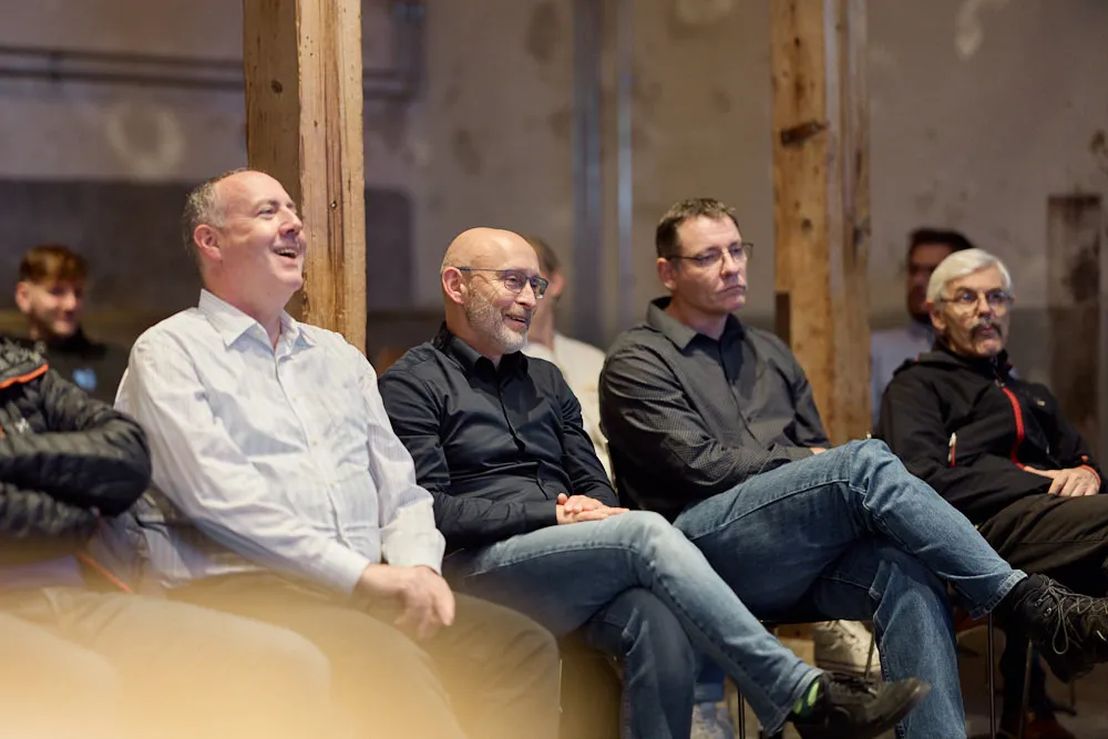 Im Fokus sind vier Männer im Publikum. Sie sitzen. Zwei Männer lächeln, die anderen zwei schauen ernst.