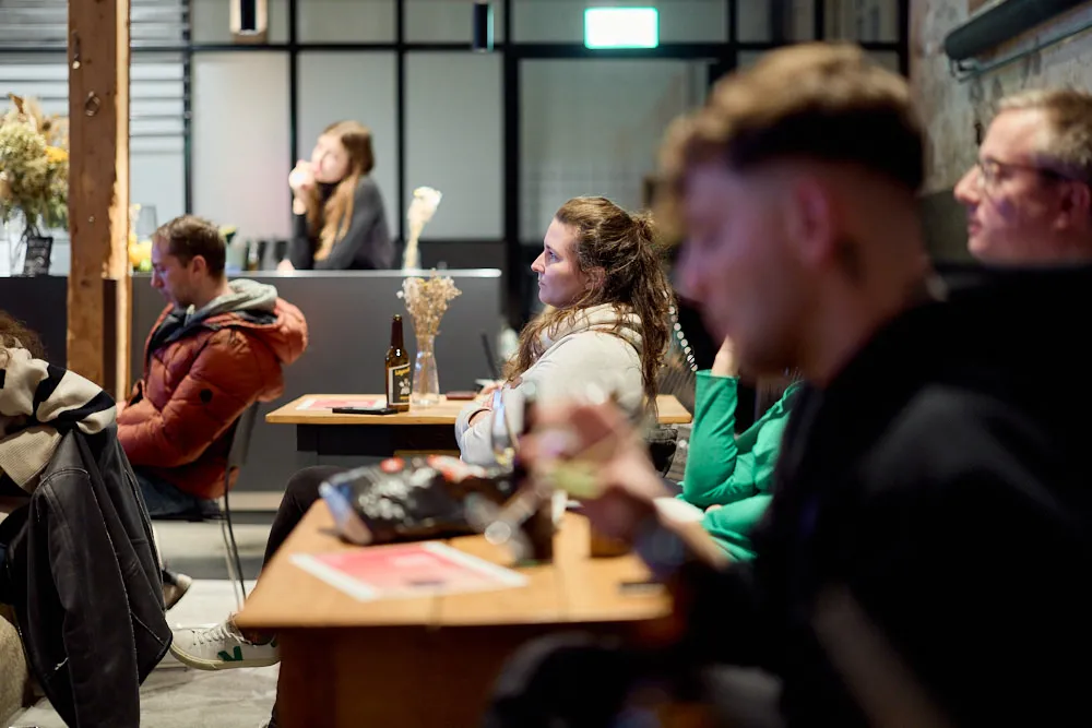 Ein Publikum sitzt einzeln vor Tischen. Auf den Tischen befinden sich Getränke und Flyer.