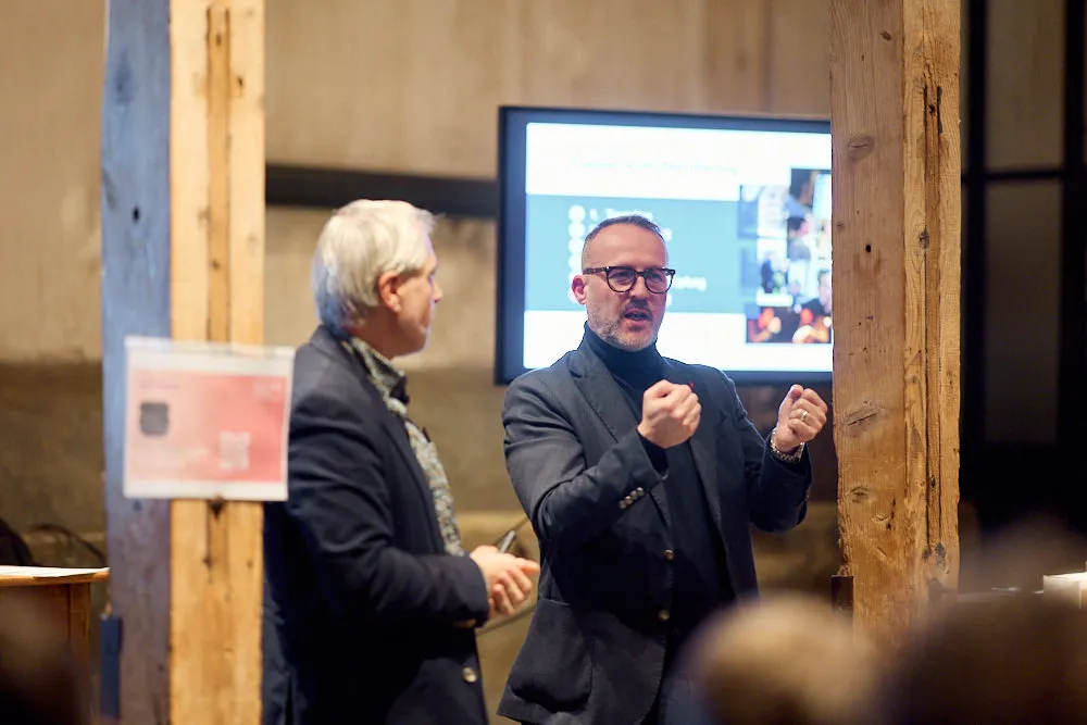 Zwei Männer sprechen vor einem Publikum in einem Raum.