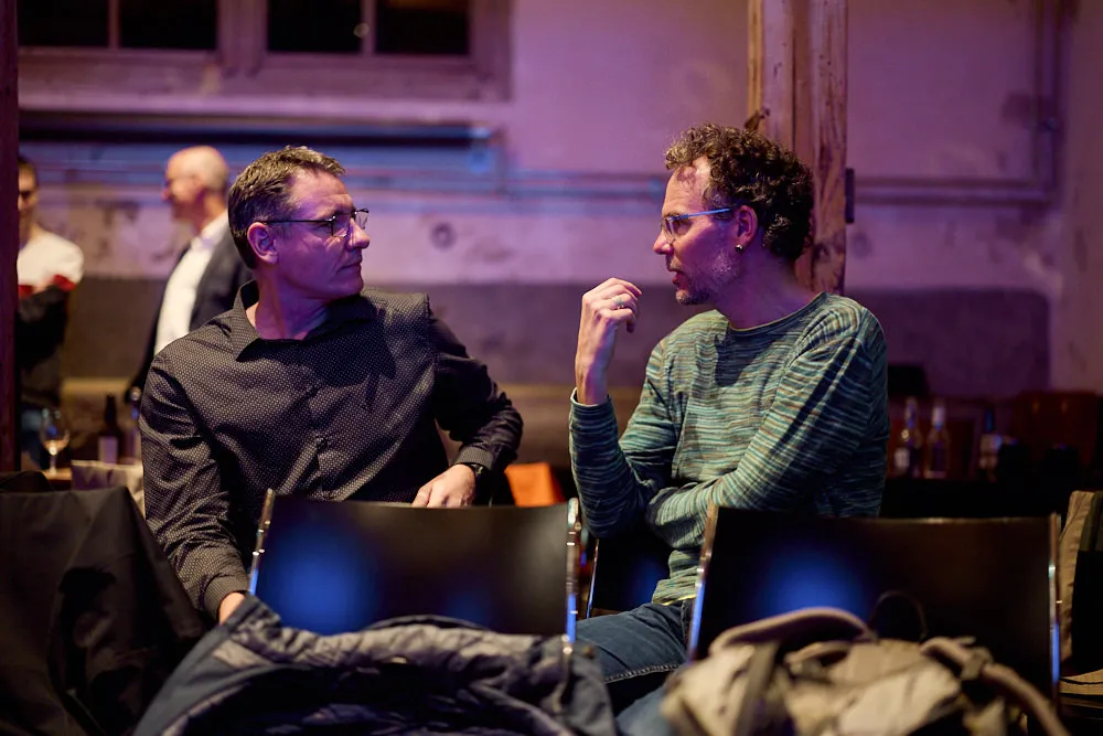 Zwei Männer unterhalten sich im Publikum auf Stühlen. Die Präsentation scheint beendet zu sein.