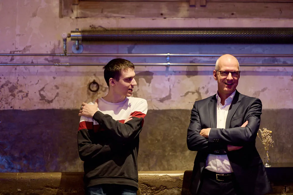 Zwei Männer stehen mit verschlossenen Armen bei einer Wand. Der eine lächelnd den anderen an. Der andere schaut lächelnd nach vorne.