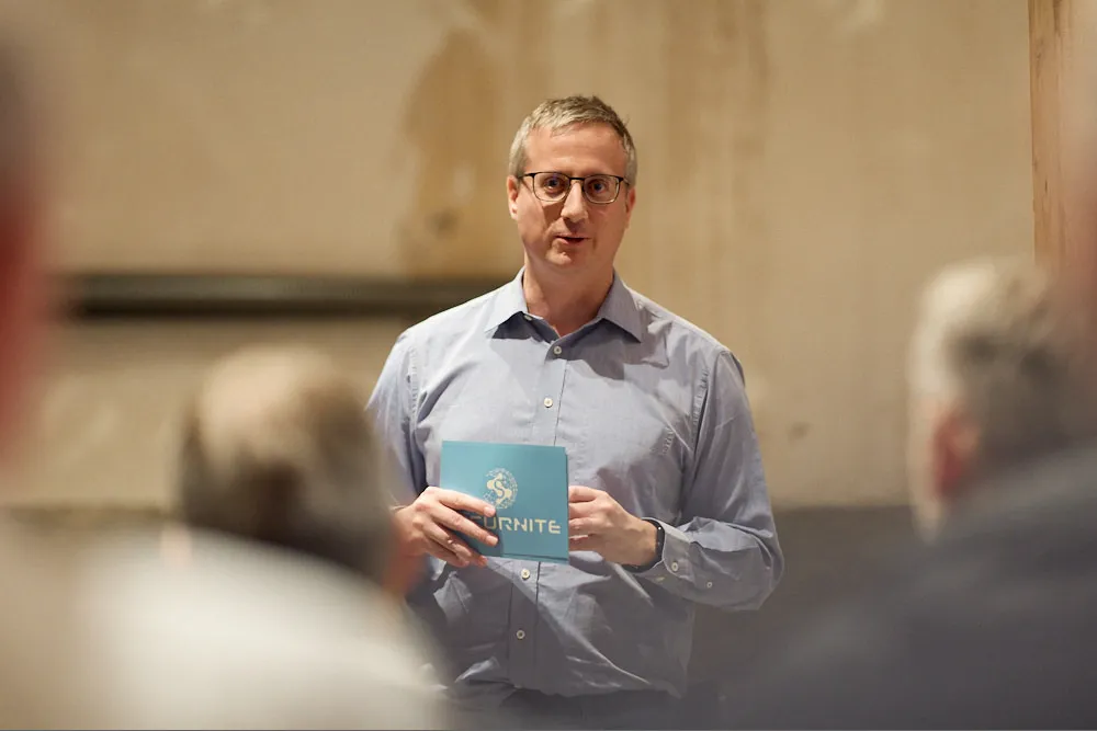 Ein Mann in Hemd spricht vor einem Publikum. Er hält eine Karte in der Hand.