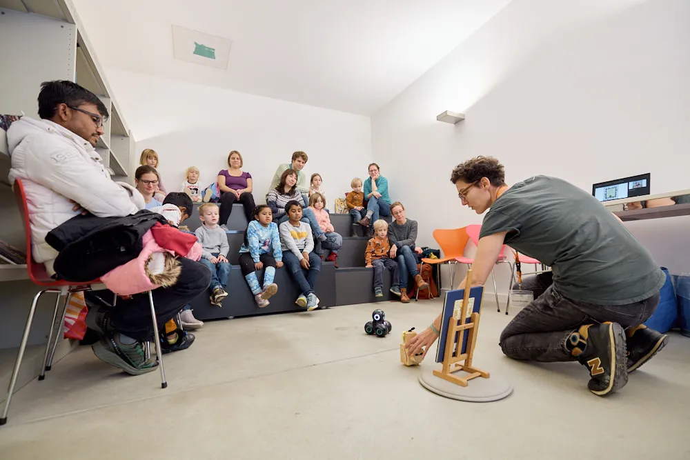 Eine Frau erzählt mit dem Buch und Robotern eine Geschichte an Kindern und Eltern auf einer kleinen Tribüne.