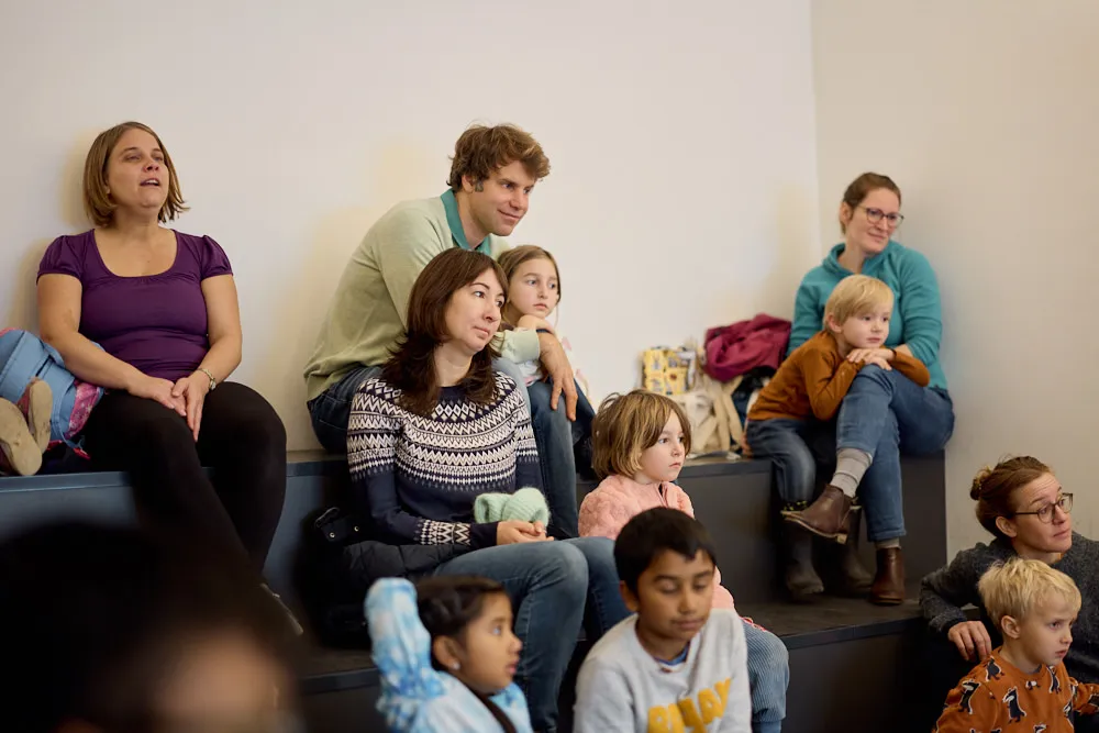Eltern und Kinder auf einer Tribüne hören gespannt zu.