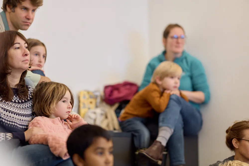 Eltern und Kinder auf einer Tribüne hören gespannt zu.