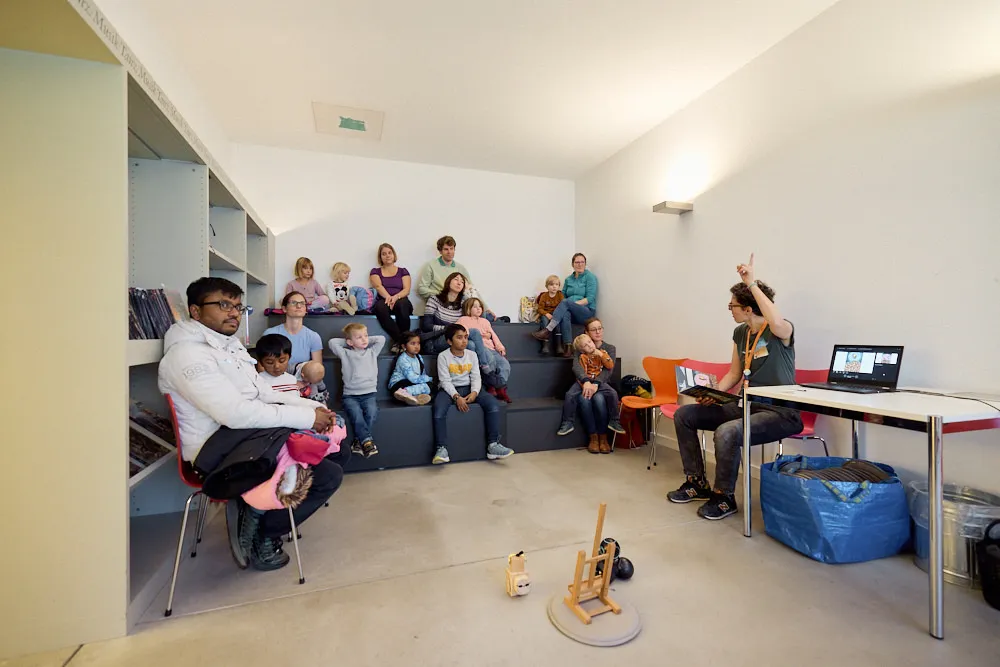 Eine Frau erzählt mit dem Buch und Robotern eine Geschichte an Kindern und Eltern auf einer kleinen Tribüne.