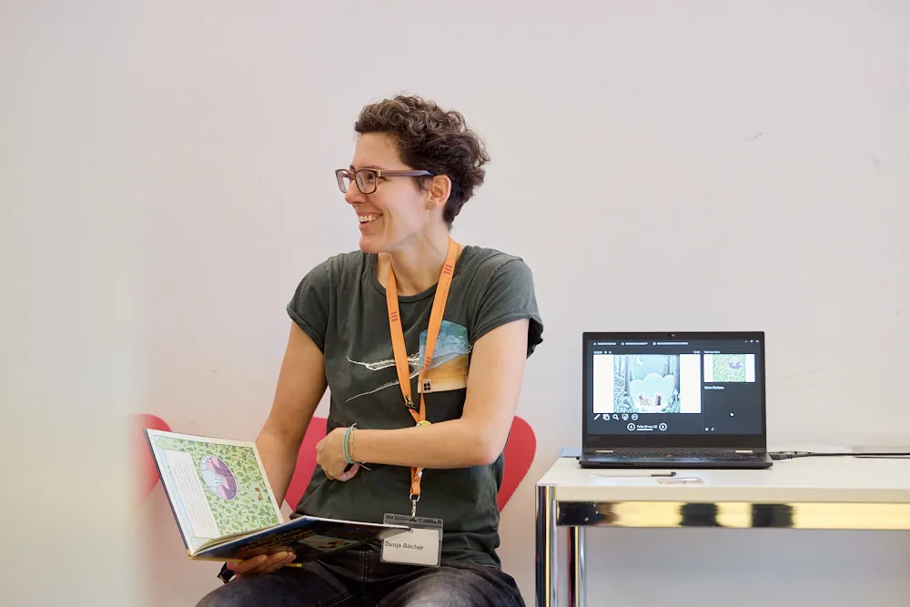 Eine Frau erzählt lächelnd aus einem Kinderbuch. Neben ihr befindet sich ein geöffneter Laptop der ein geöffnetes Programm zeigt.