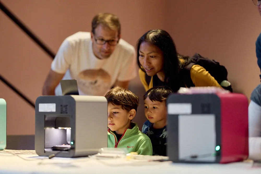 Ein Mann und eine Frau schauen mit zwei Knaben in den würfelförmigen 3D-Drucker.