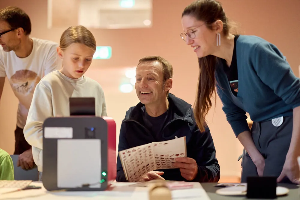 Ein Mann hält ein Heft in der Hand und schaut mit einem Mädchen und einer Frau in den 3D-Drucker.
