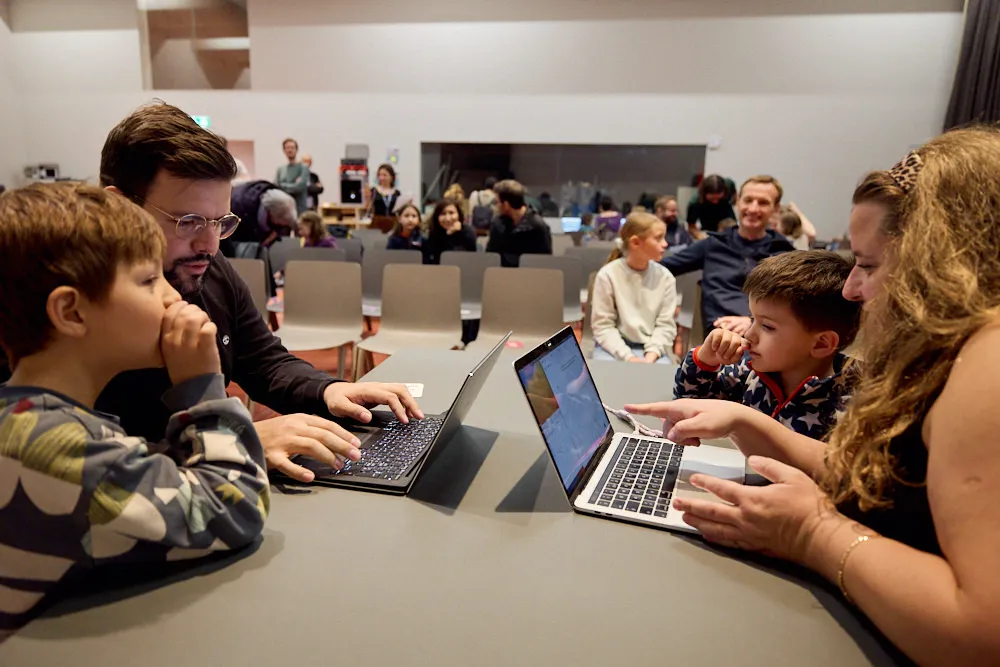 Ein Mann mit Kind und eine Frau mit Kind schauen alle jeweils in einen Laptop. Sie befinden sich jeweils gegenüber.