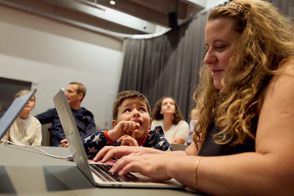 Eine Frau ist am Laptop. Ein Kind nebenan schaut ihr gespannt zu.