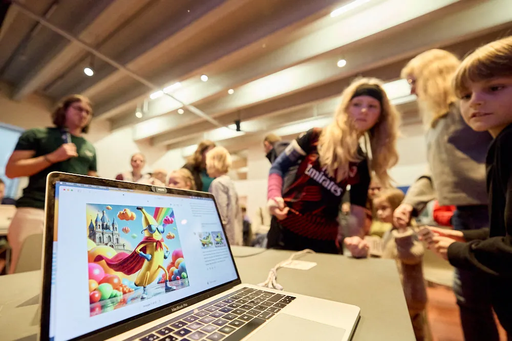 Kinder schauen interessiert in einen Laptop. Im Laptop wird ein lustiges Bild gezeigt. Auf dem Bild ist eine Banane in Superheldengewand auf farbigen Wolken und Schloss im Hintergrund.