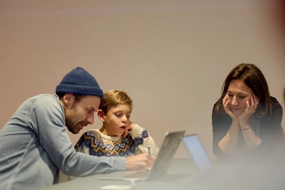 Ein Mann mit einem Jungen schauen gespannt in einen Laptop. Eine Frau sitzt neben ihnen.