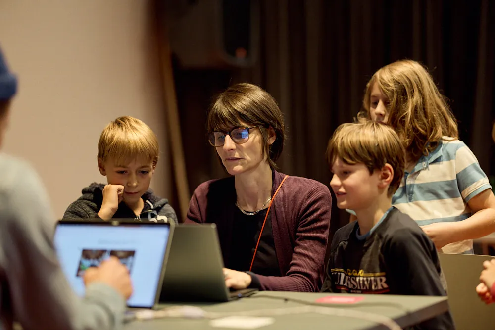Eine Frau bedient einen Laptop. Neben und hinter ihr sind drei Kinder. Sie schauen der Frau zu.