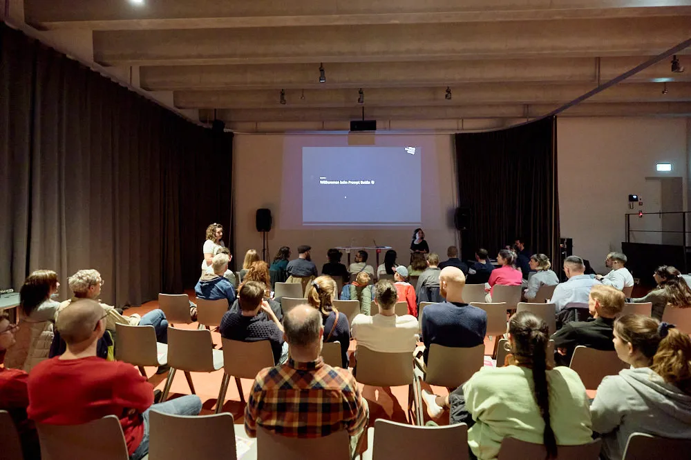 Ein Publikum sitzt auf Stühlen in einem Raum und schaut auf eine Bühne. Hinter der Bühne wird eine Folie projiziert.