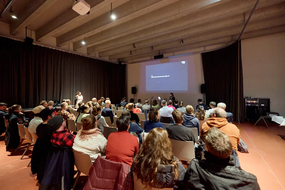 Ein Publikum sitzt auf Stühlen in einem Raum und schaut auf eine Bühne. Hinter der Bühne wird eine Folie projiziert.