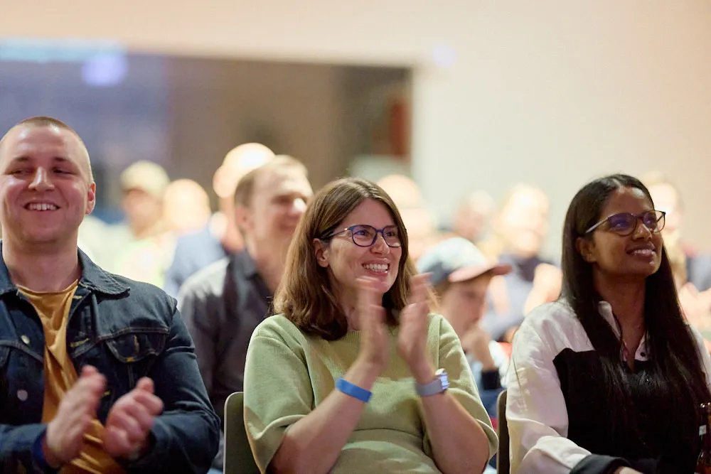 Im Fokus sind ein Mann und zwei Frauen im Publikum. Sie lächeln und klatschen.