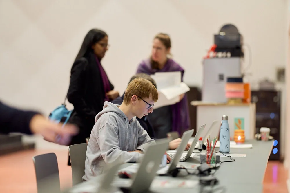 Im Fokus ist ein junger Mensch an einem Laptop. Im Hintergrunde befinden sich zwei Frauen.