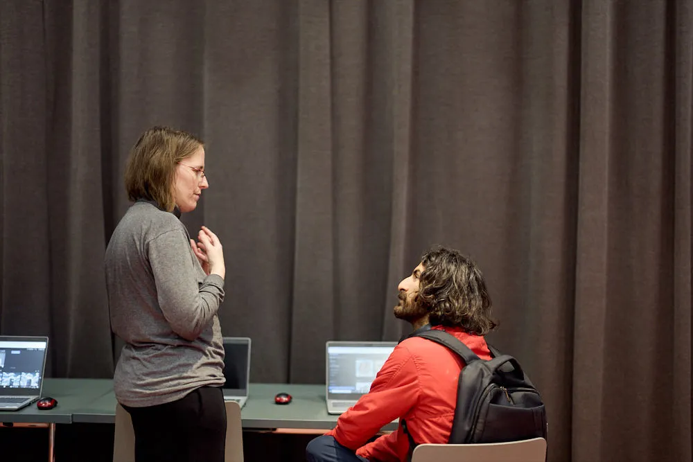 Ein Mann sitzt vor einem Laptop. Neben ihm steht eine Frau. Sie schauen sich gegenseitig an und sprechen miteinander.