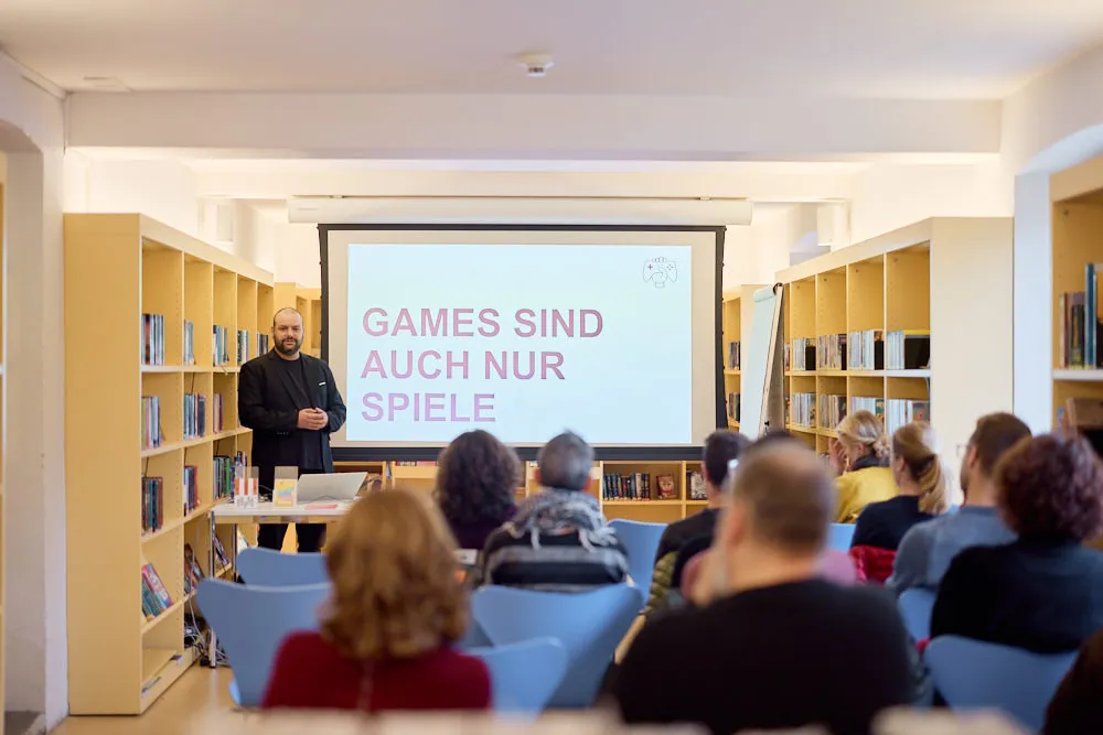 Ein Mann hält in einer Bibliothek eine Präsentation vor einer Leinwand vor einem Publikum. Auf der Leinwand wird seine Präsentation projiziert.