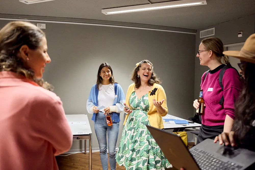 Eine Frau hält stehend und lachend eine Begrüssung. Frauen stehen um sie herum.