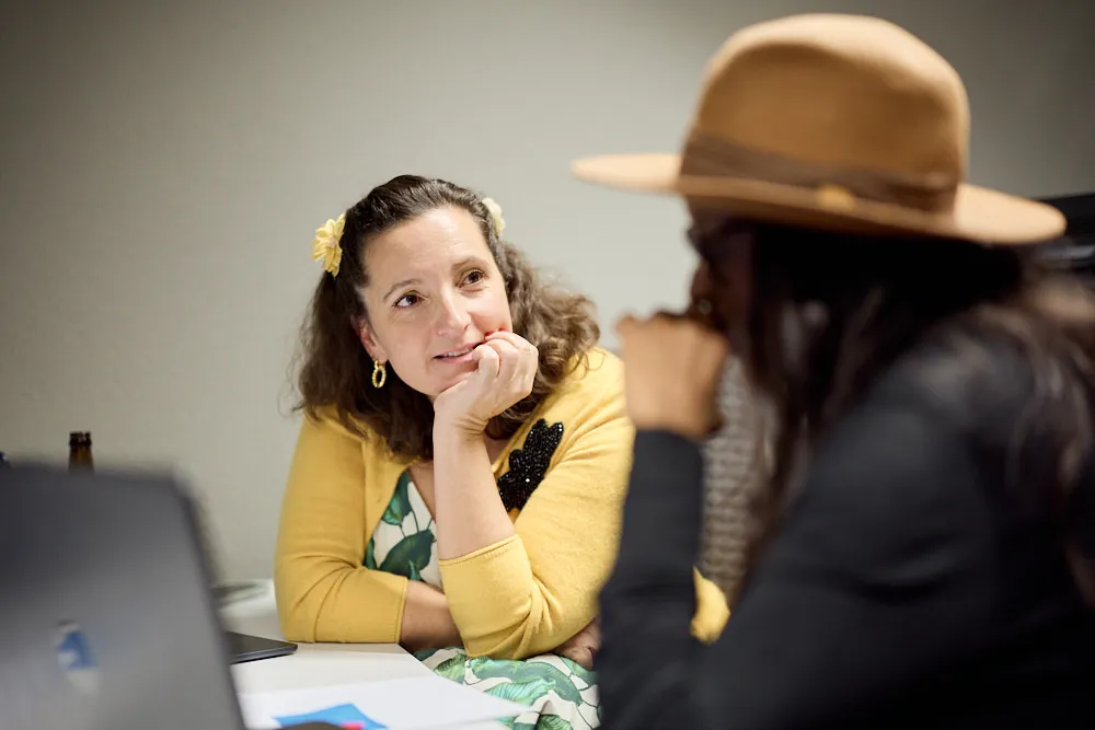 Zwei Frauen an einem Tisch sprechen miteinander. Die eine Frau schaut die andere Frau an. Die andere Frau schaut in ihrem Laptop.