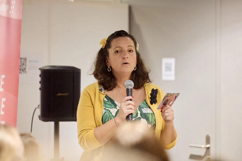 Eine Frau hält in einer Hand eine Karte und in der anderen Hand ein Mikrofon. Sie spricht ins Mikrofon vor einem Publikum.