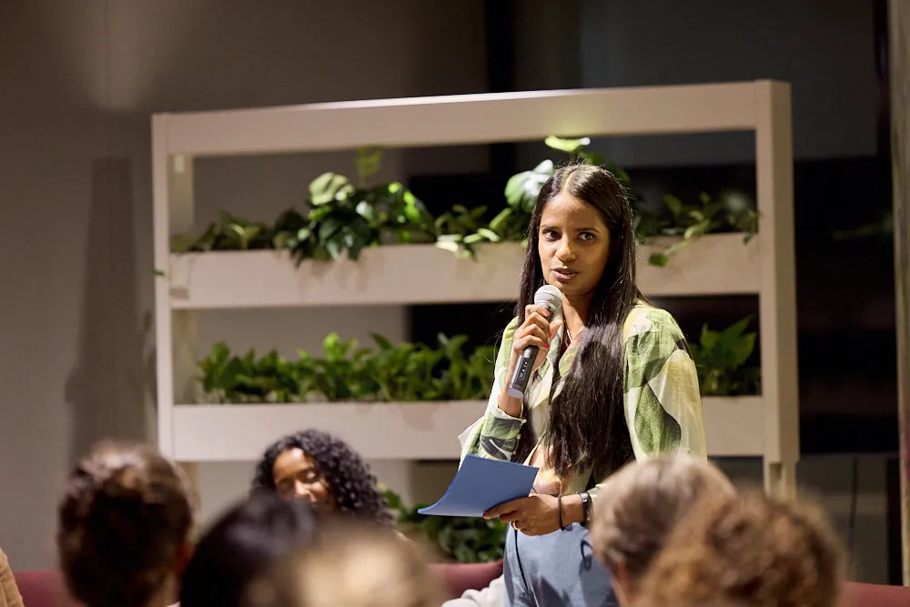 Eine Frau hält in einer Hand eine Karte und in der anderen Hand ein Mikrofon. Sie spricht ins Mikrofon vor einem Publikum.