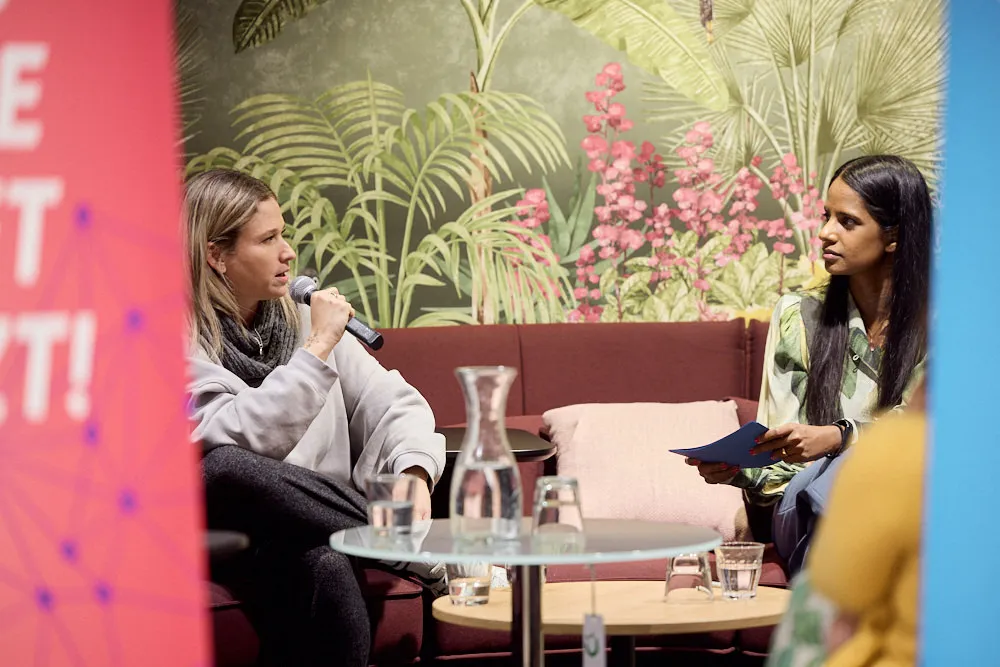 Zwei Frauen auf dem Sofa schauen sich gegenseitig an. Eine Frau spricht in ein Mikrofon zu der Frau links von ihr.