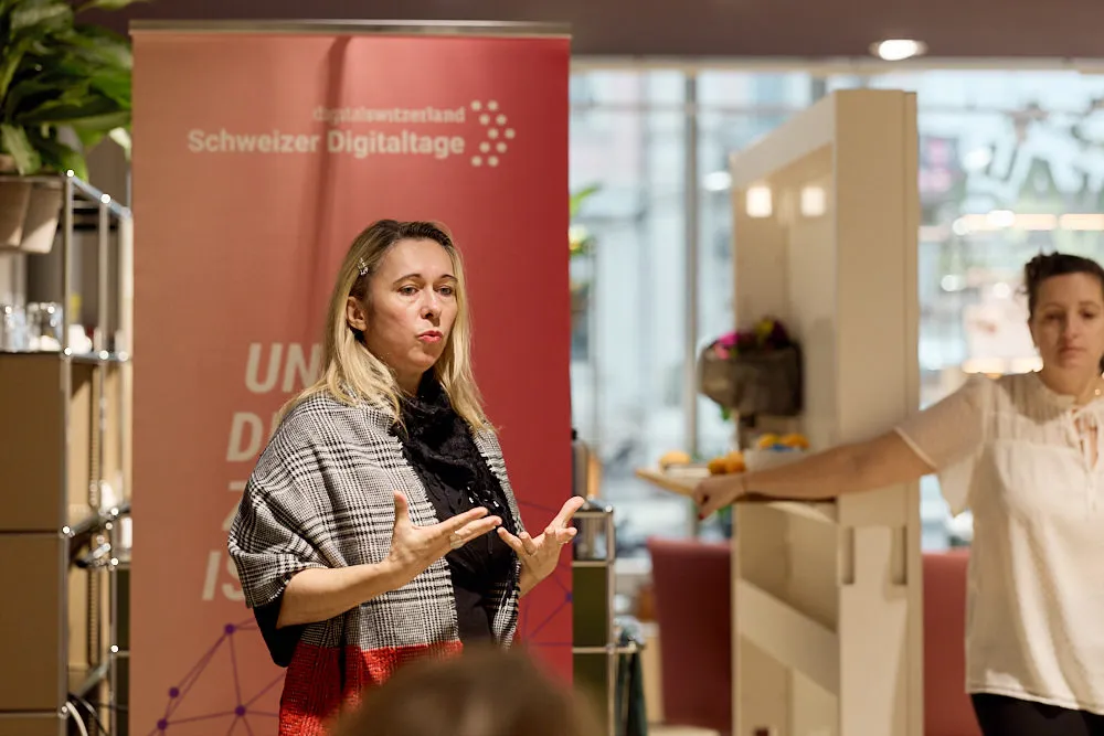 Eine Frau hält eine Präsentation. Hinter ihr im Hintergrund befindet sich ein Roll-Up und eine Frau.