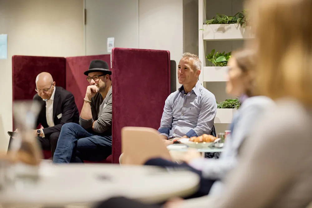 Menschen an Tischchen mit Brötchen und Menschen in Nischen hören gespannt zu.