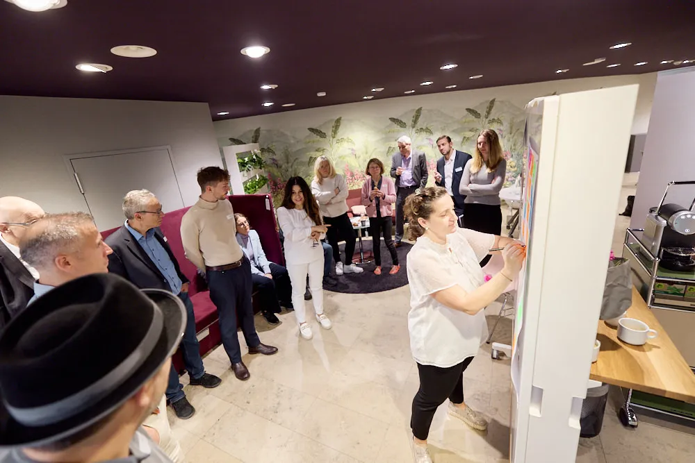 Eine Frau schreibt etwas auf ein Post-It auf eine Wand. Menschen stehen hinter ihr und schauen ihr zu.