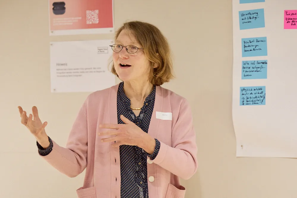 Eine Frau erzählt etwas vor einer Wand mit Post-Its. sie gestikuliert mit den Händen.