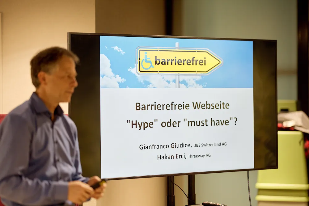 Ein Mann hält eine Präsentation vor einem Bildschirm. Auf dem Bildschirm steht der Titel Barrierefreie Website Hype oder Must-Have?