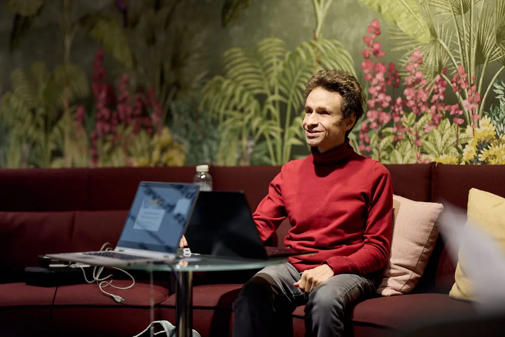 Ein Mann sitzt auf einem langen Sofa vor einem Tisch mit zwei Laptops. Er schaut nach vorne und lächelt.