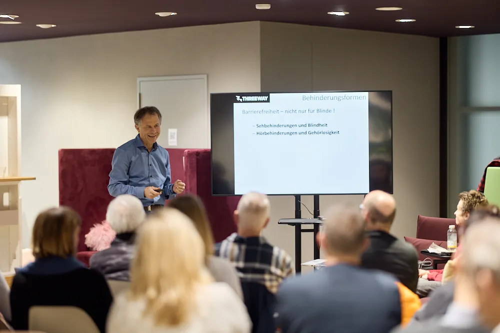 Ein Mann hält eine Präsentation vor einem Bildschirm. Ein Publikum hört ihm zu.