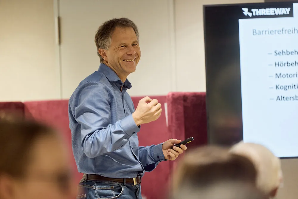 Ein Mann hält lächelnd eine Präsentation vor einem Bildschirm. Auf dem Bildschirm steht der Titel Barrierefreie Website Hype oder Must-Have?Ein Mann hält eine Präsentation vor einem Bildschirm.