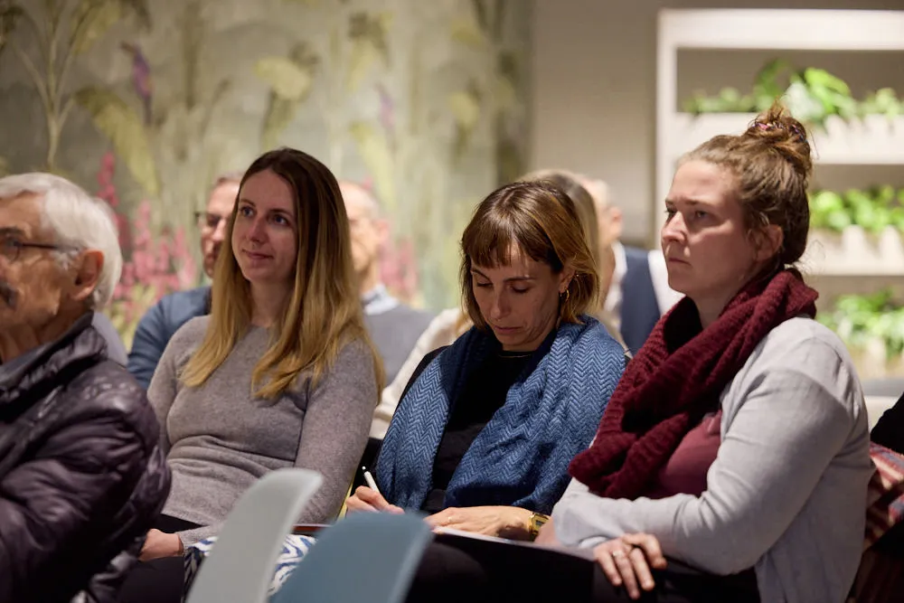 Im Fokus sind drei Frauen im sitzenden Publikum. Die Frau in der Mitte schreibt etwas auf.