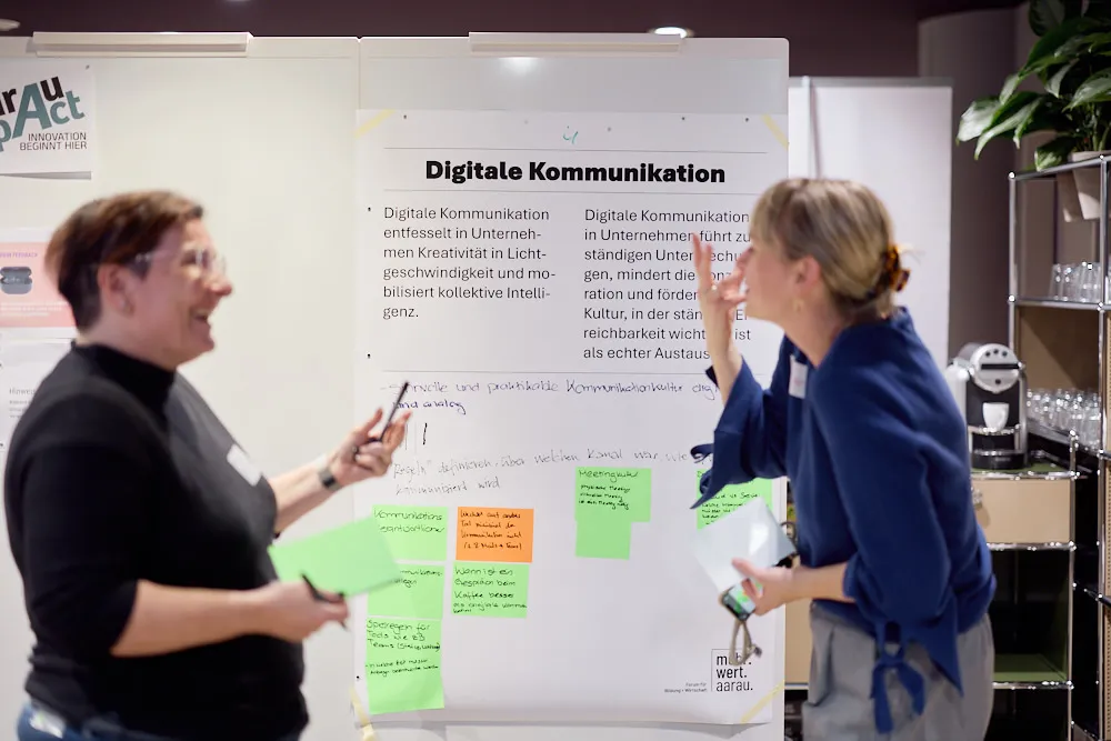 Zwei Frauen sprechen aufgeregt und lächelnd vor einem Plakat mit dem Titel Digitale Kommunikation miteinander.