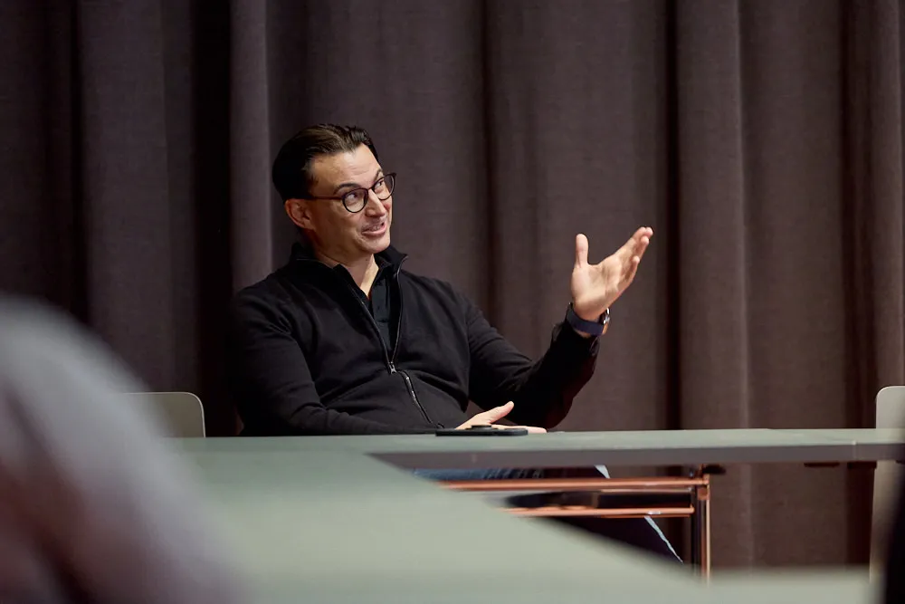 Ein sitzender Mann an einem Tisch erzählt etwas und gestikuliert dabei. Er lächelt.