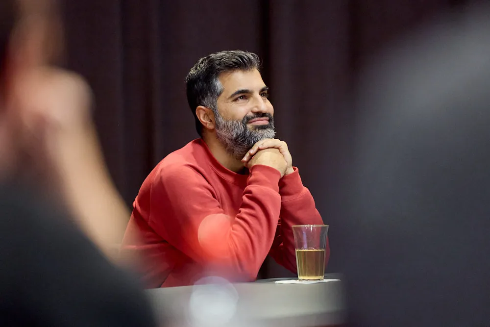 Ein Mann sitzt und stützt seinen Kopf auf den Händen. Er hört gespannt und lächelnd zu. Vor ihm steht ein transparenter Becher mit einem Getränk.