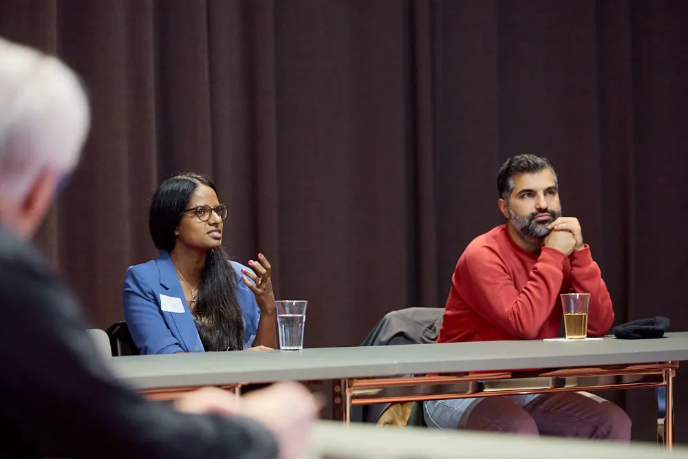 Im Fokus sind eine Frau und ein Mann an einem Tisch. Sie sitzen. Die Frau spricht und gestikuliert mit einer Hand.
